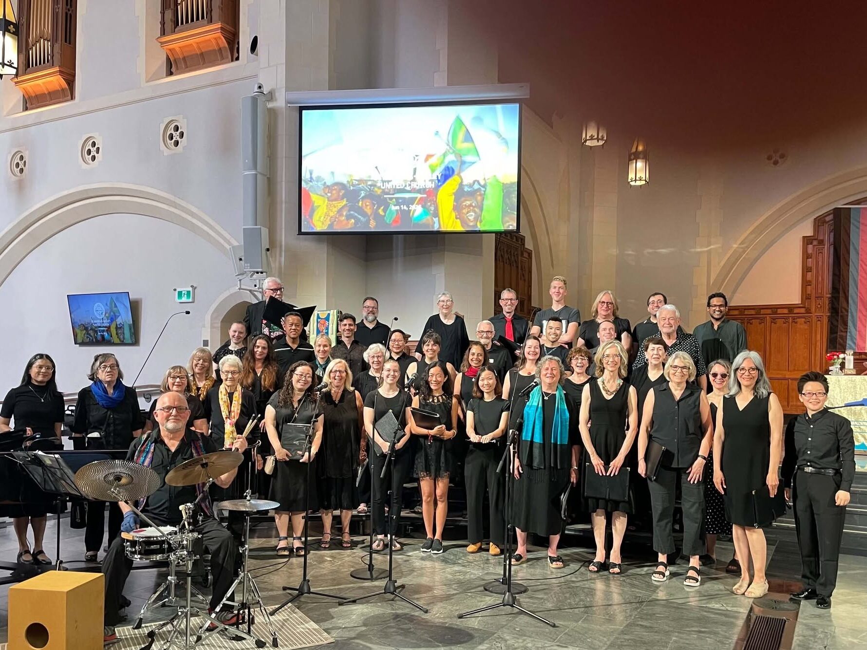 A happy and smiling choir, the West End Singers, stands at the front of St. Andrew's Wesley United Church. There is a drummer and drum kit in the foreground.